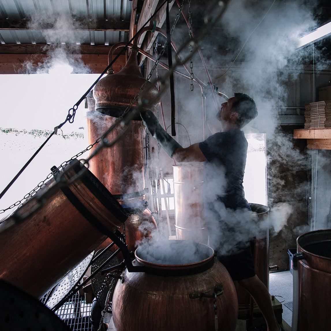 Person operating copper distillation equipment amid steam inside a distillery