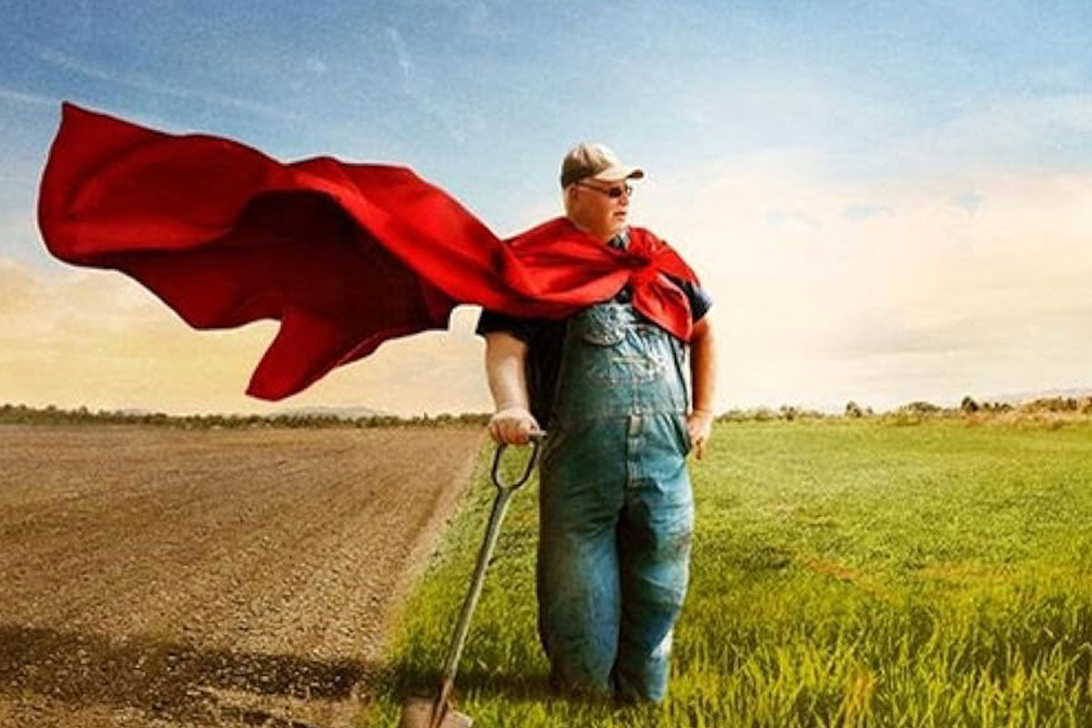 Farmer wearing a red superhero cape standing with a shovel between a plowed field and green pasture under a pale sky