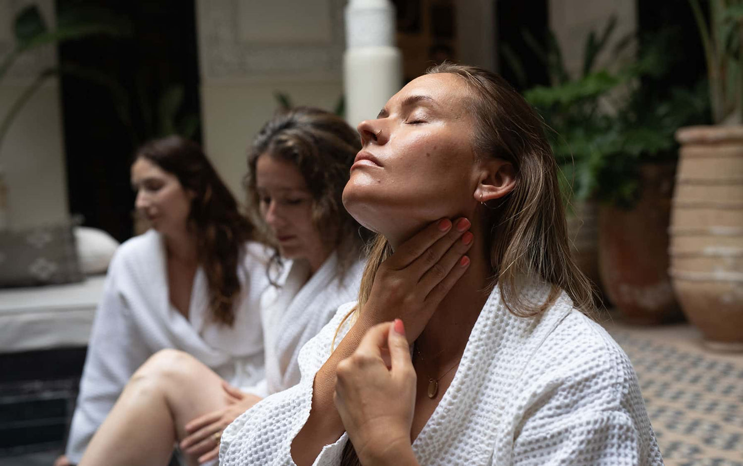 Woman in foreground with eyes closed touching her neck, two women in robes seated behind in a sunlit spa courtyard