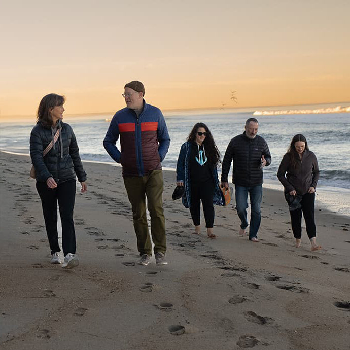 Five people walking along a beach at sunset, some barefoot, wearing jackets and leaving footprints in the sand