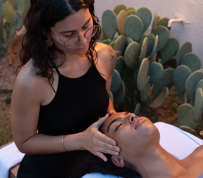 Esthetician wearing glasses gives a facial massage to a reclining client outdoors near prickly pear cactus