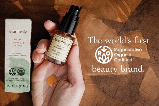Hand holding Balm of Gilead Elixir bottle beside its box, pump dispenser and product droplet on a wood background