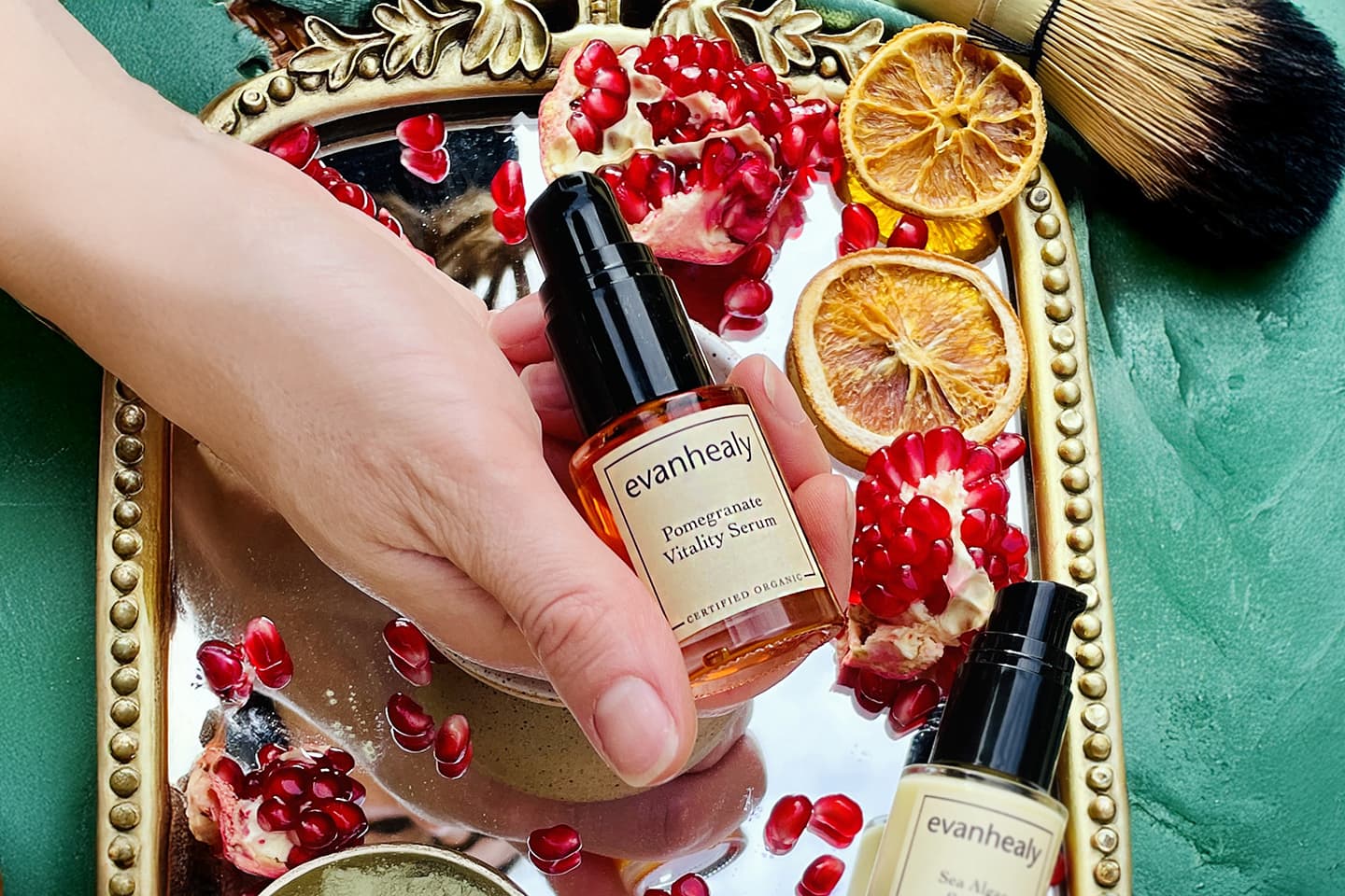 Close-up of evanhealy Pomegranate Vitality Serum showcasing its amber bottle held over pomegranate seeds and dried citrus