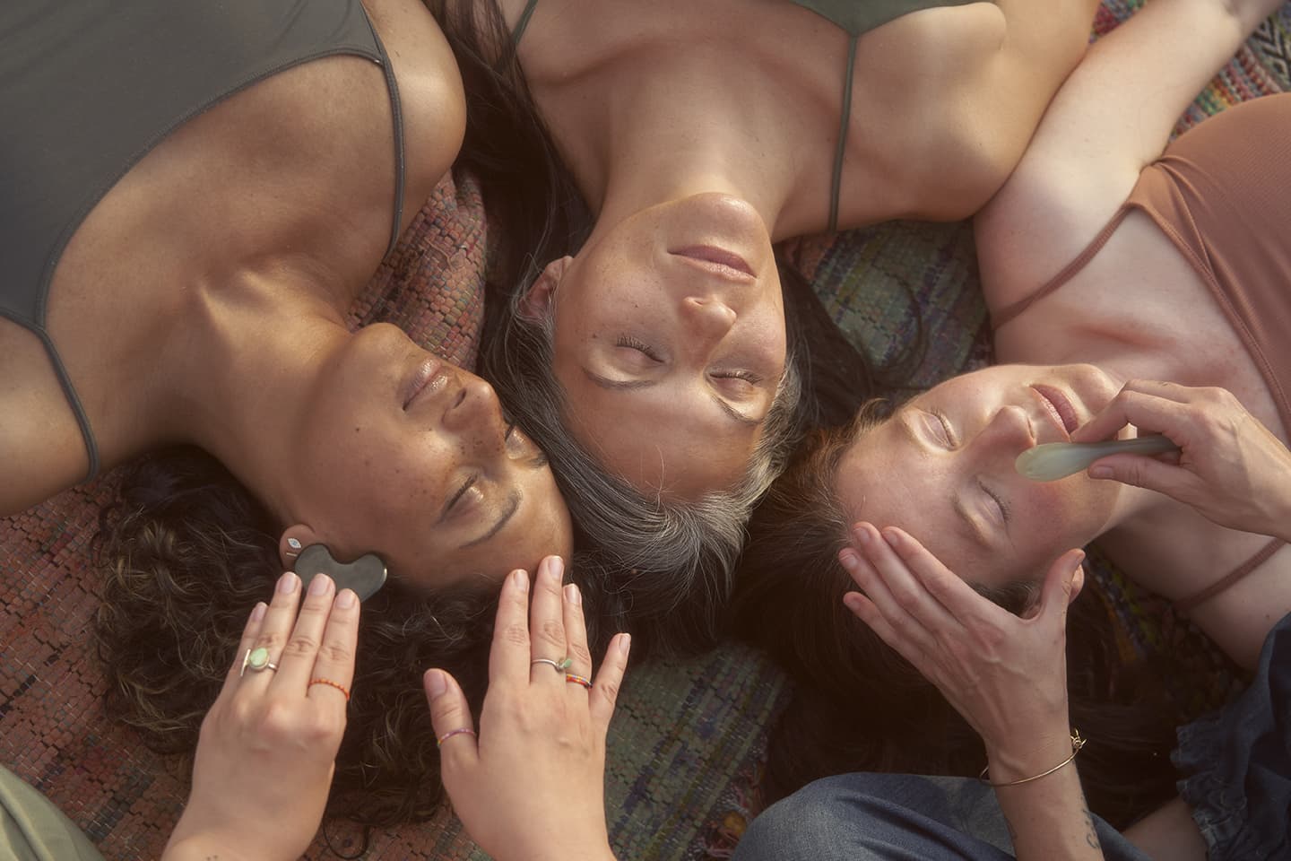 Three women lying on a patterned blanket while receiving facial massages with stone tools