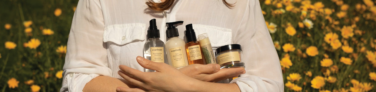 Person holding five Evan Healy skincare bottles and jars in a sunlit field of yellow flowers