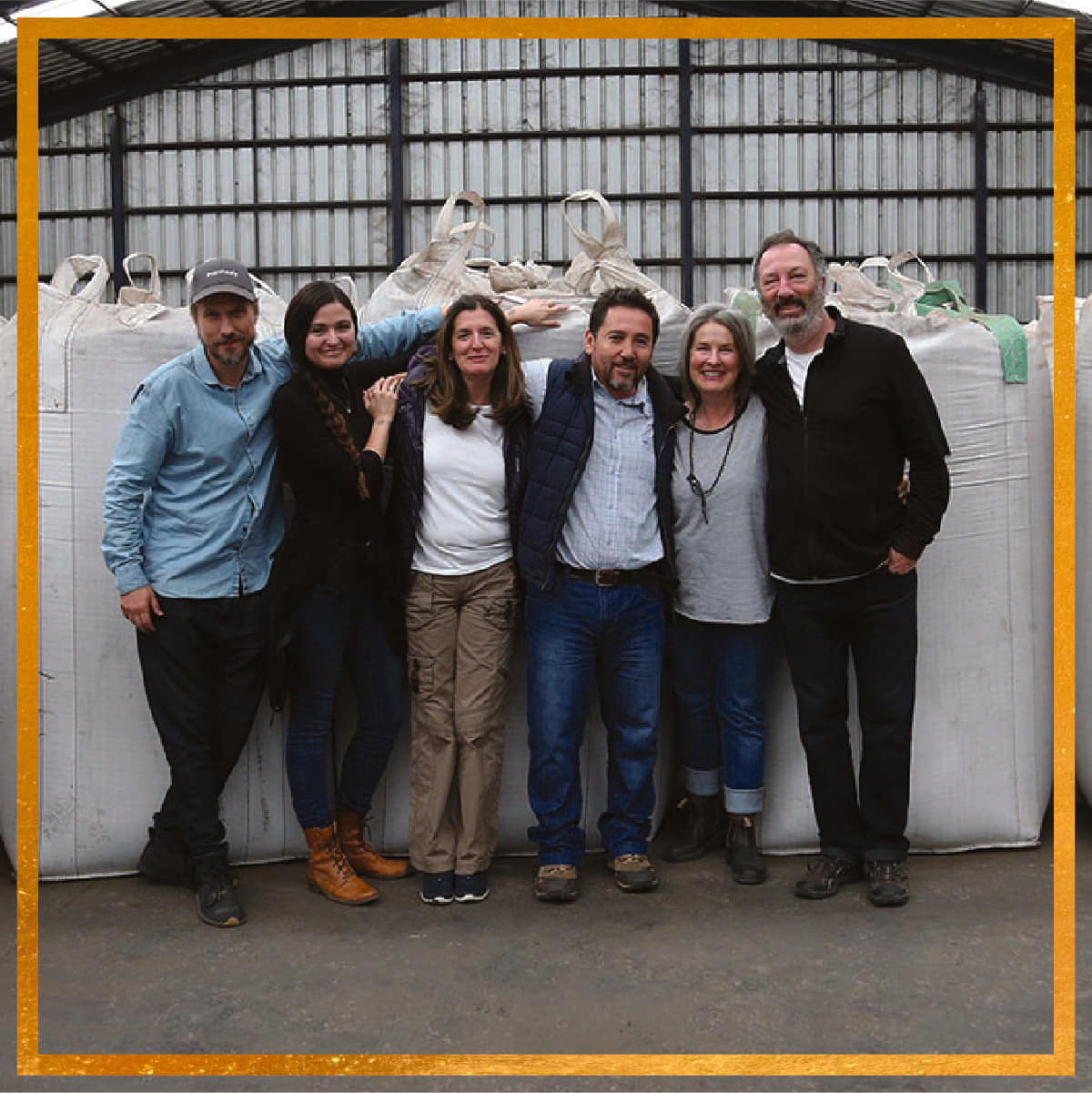 Six people standing arm-in-arm in a warehouse in front of large white bulk bags