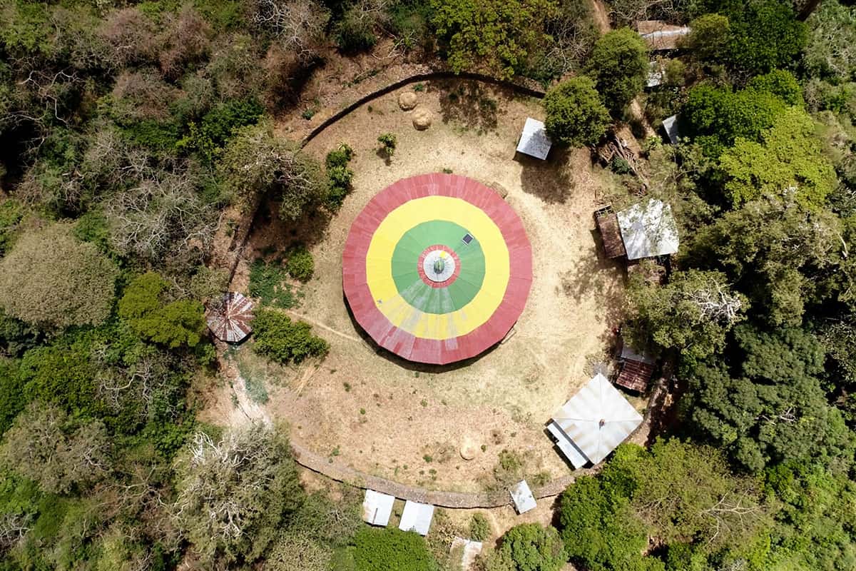 Aerial view of a circular church with a red, yellow, and green striped roof surrounded by trees in an Ethiopian church forest