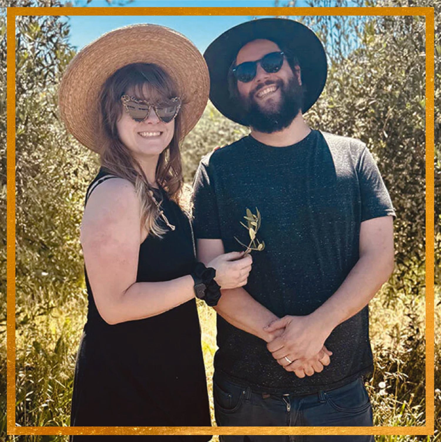 Two people smiling in a sunlit olive grove, woman wearing a straw hat holding an olive sprig