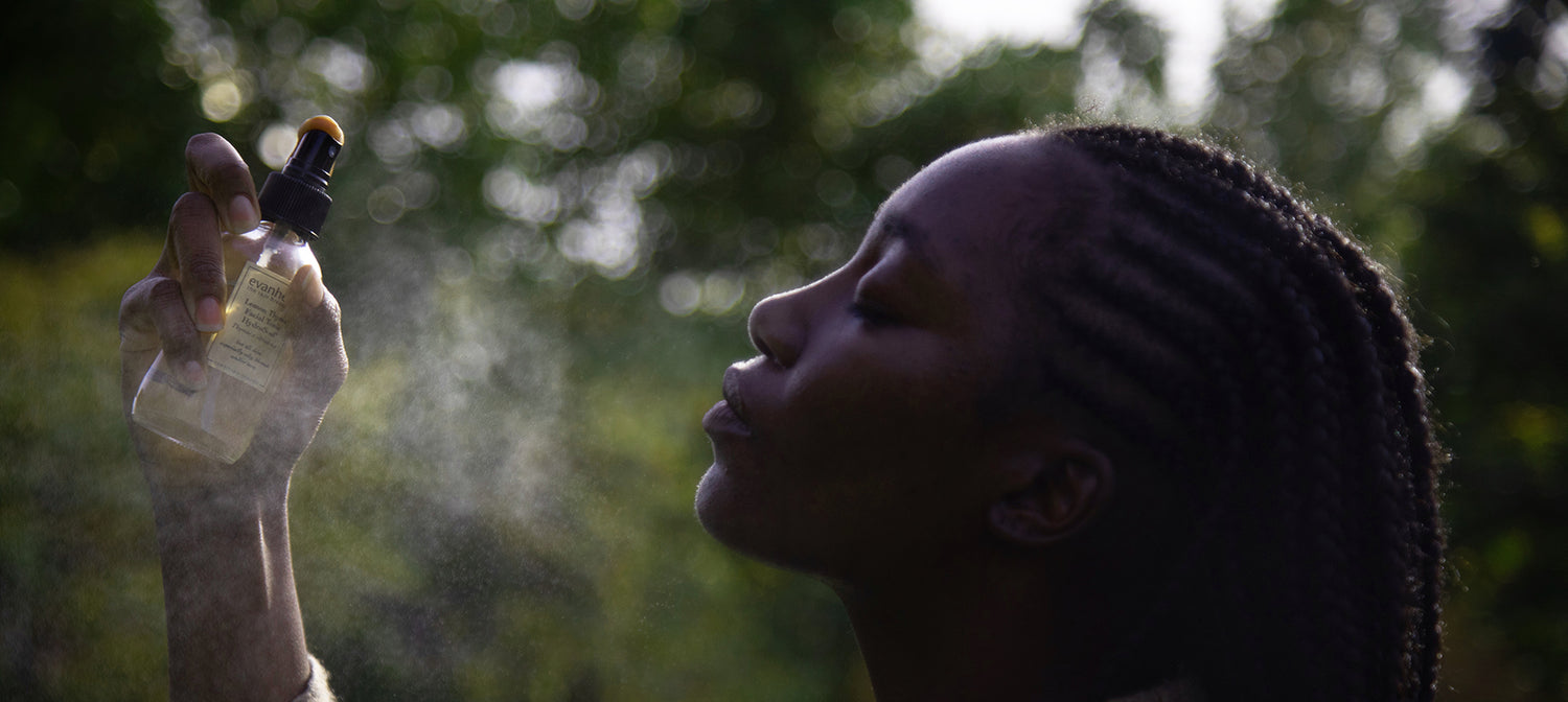 Person with braided hair spritzing a facial mist from a glass spray bottle outdoors with sunlight filtering through trees and visible mist