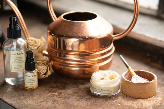 Evanhealy skincare bottles and an open cream jar displayed beside a copper watering can and ceramic bowl on a rustic windowsill