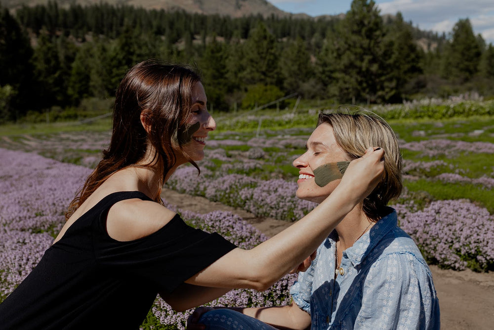 Two women applying green clay face mask to each other while seated in a lavender field