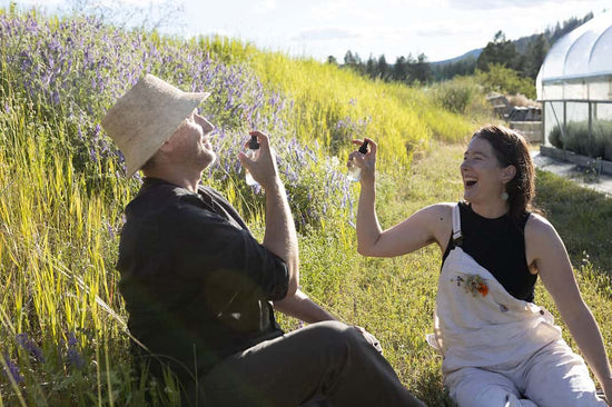 Two people laughing and spraying facial mists while seated in a sunlit lavender field with a greenhouse in the background