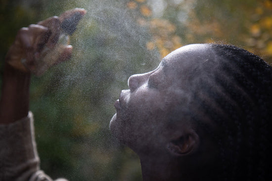 Close-up profile of a woman misting her face with a facial spray against sunlit, blurred greenery