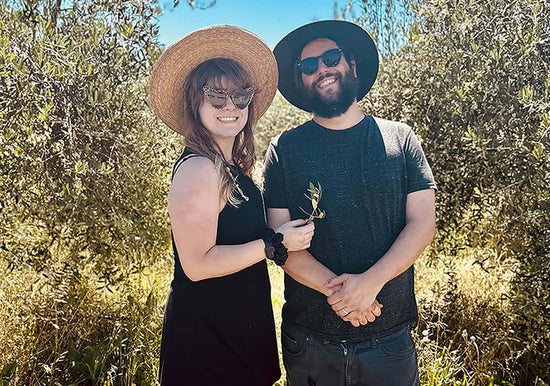 Two stewards smiling in a sunlit olive grove, woman in straw hat holding an olive branch, man in black hat and sunglasses