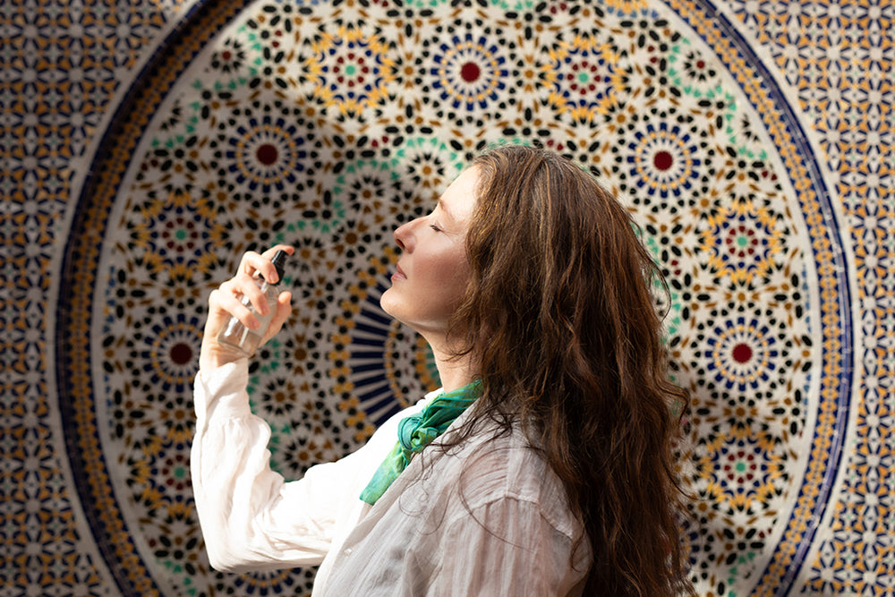 Woman misting her face with facial hydrosol in front of a colorful Moroccan mosaic wall