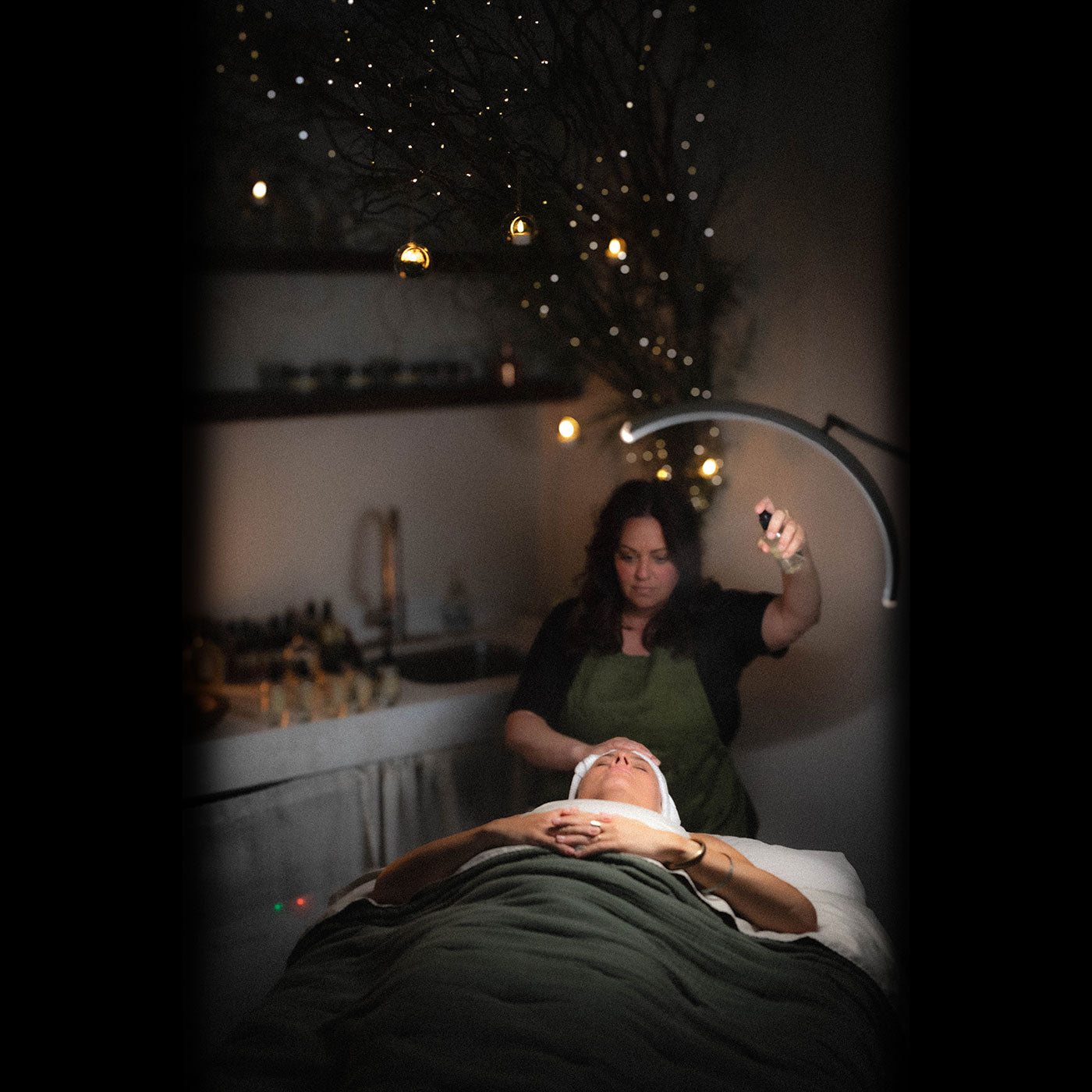 Esthetician misting a client during a restorative facial in a dim, cozy treatment room with warm string lights