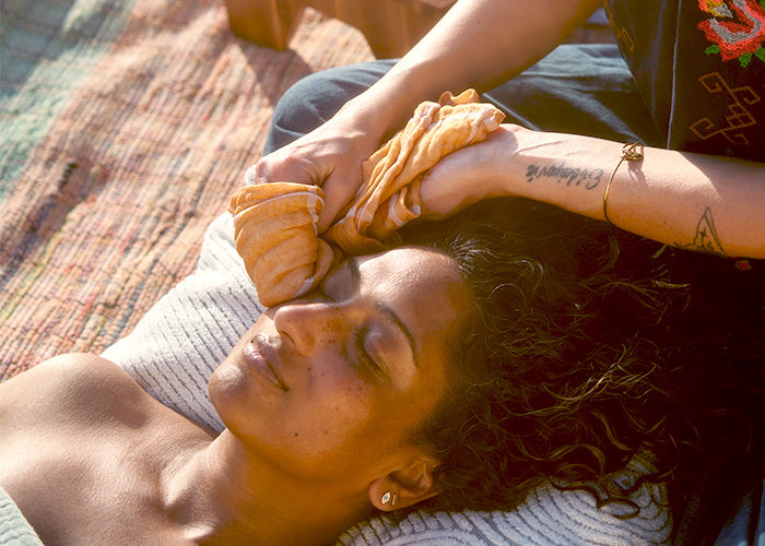 Woman lying on a blanket outdoors receiving a warm facial compress from a practitioner with tattooed hands