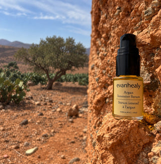 Front view of Argan Intensive Serum showcasing its amber bottle and black pump on a rocky ledge with an argan grove