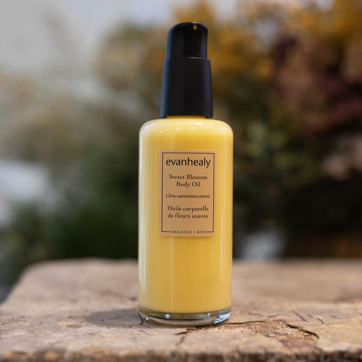 Front view of Sweet Blossom Body Oil showcasing its yellow oil in a pump bottle with a blurred natural background