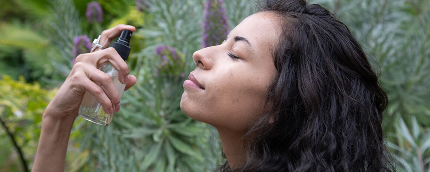 Woman with eyes closed spraying a facial mist from a clear pump bottle in a green garden setting