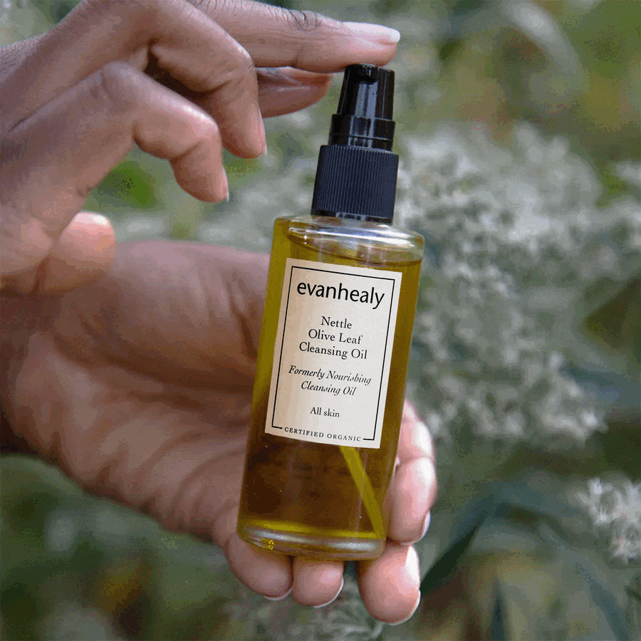 Close-up of Nettle Olive Leaf Cleansing Oil bottle with pump and Certified Organic label held against a leafy outdoor background