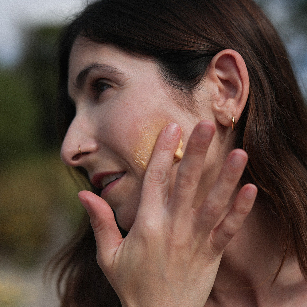 Woman applying Whipped Manuka Honey Cleansing Mask to her cheek outdoors