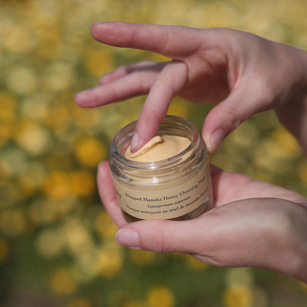 Close-up of Whipped Manuka Honey Cleansing Mask showing whipped texture in an open jar with hands against yellow flowers