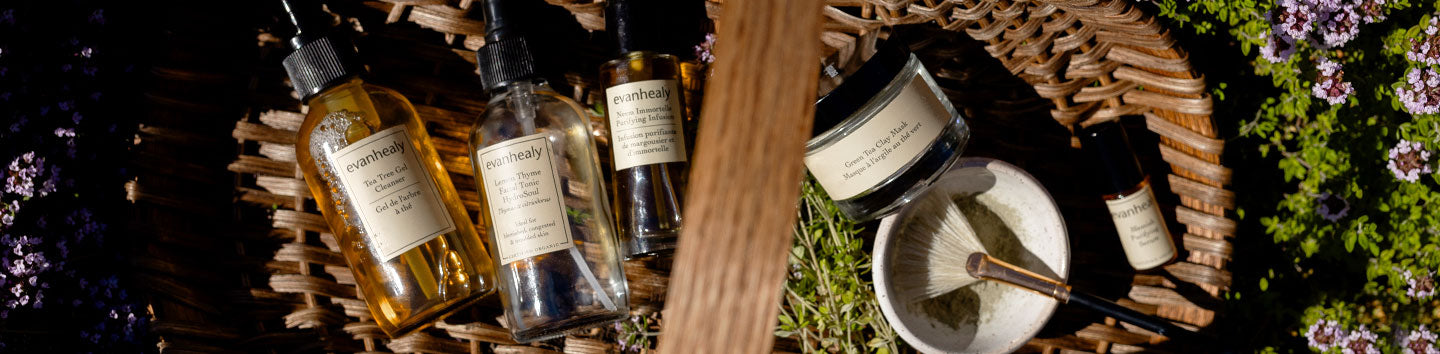 Evanhealy purifying skincare bottles and a jar arranged in a wicker basket with a mixing bowl and brush among lavender and greenery