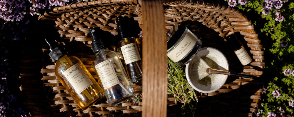Wicker basket with evanhealy skincare bottles, jars, a mixing bowl and brush arranged among small purple flowers