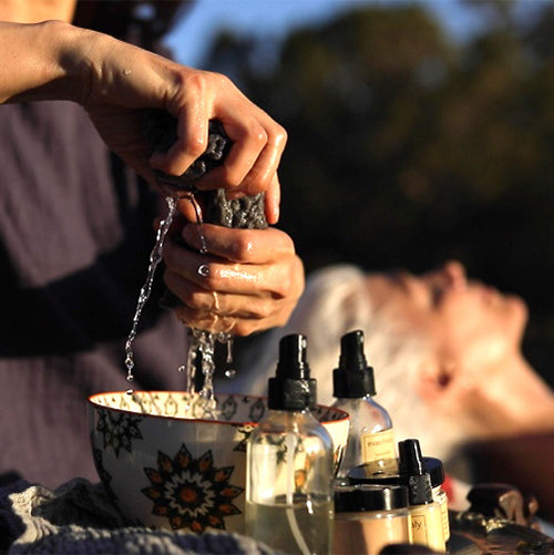 Esthetician squeezing a herbal compress over a bowl with skincare bottles in foreground and a reclining client blurred behind