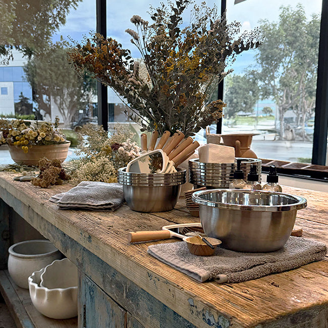 Guided facial workshop setup with stainless bowls, wood tools, towels, product bottles, and dried flowers on a rustic table