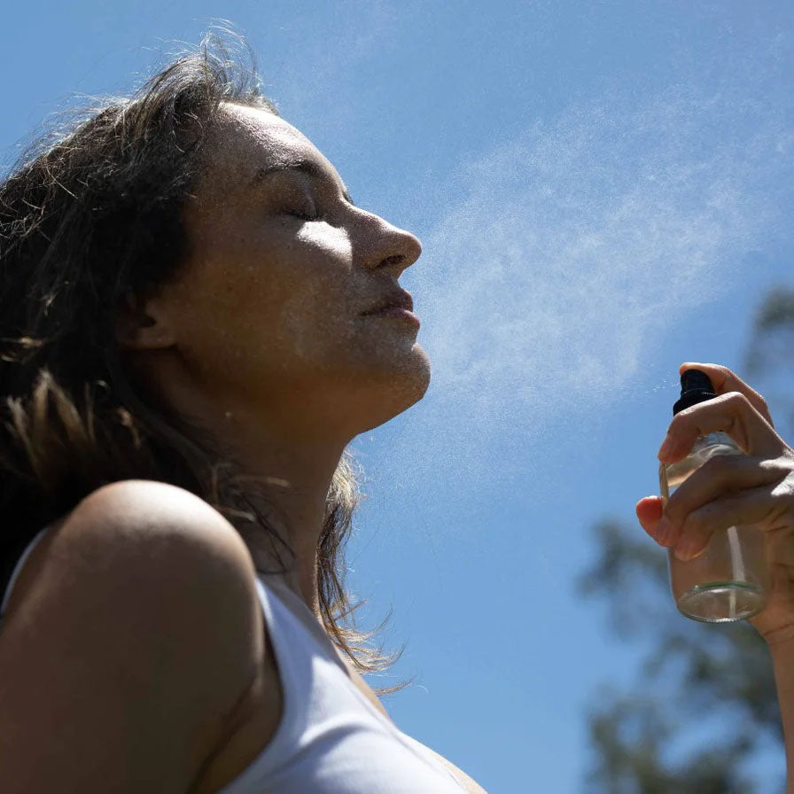 Woman spraying a facial mist from a glass spray bottle onto her face outdoors against a clear blue sky