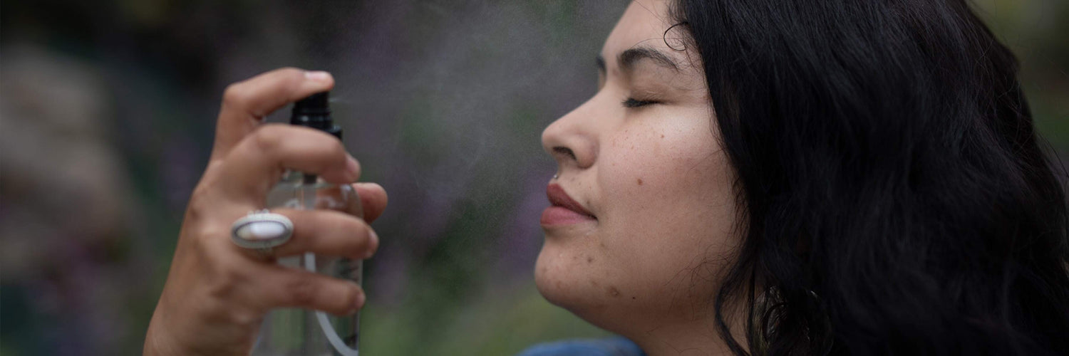 Profile of a woman with blemished skin spraying facial mist with eyes closed against a blurred outdoor background