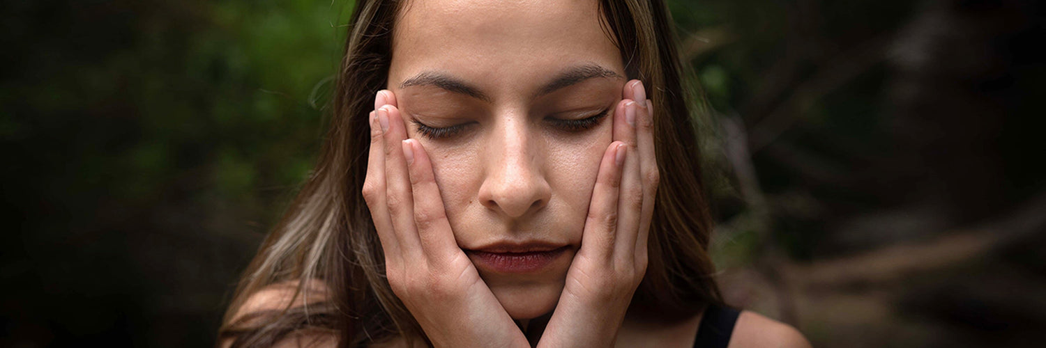 Young woman with eyes closed gently cupping her face with both hands against a blurred outdoor background