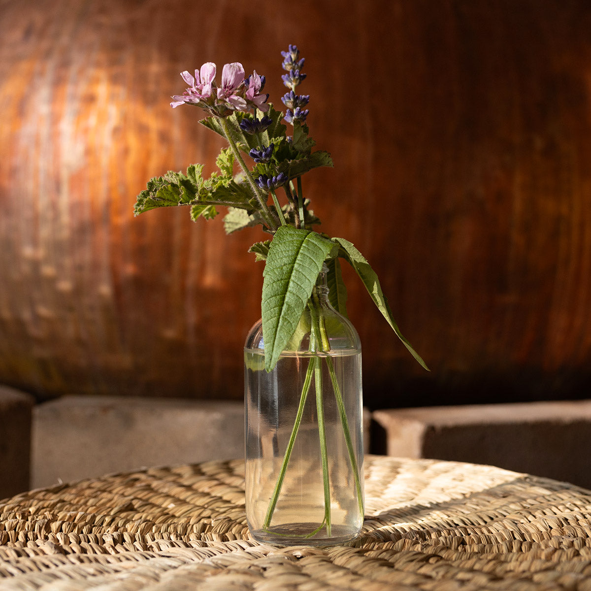 Rose geranium and purple flowers arranged in a glass vase on a woven table