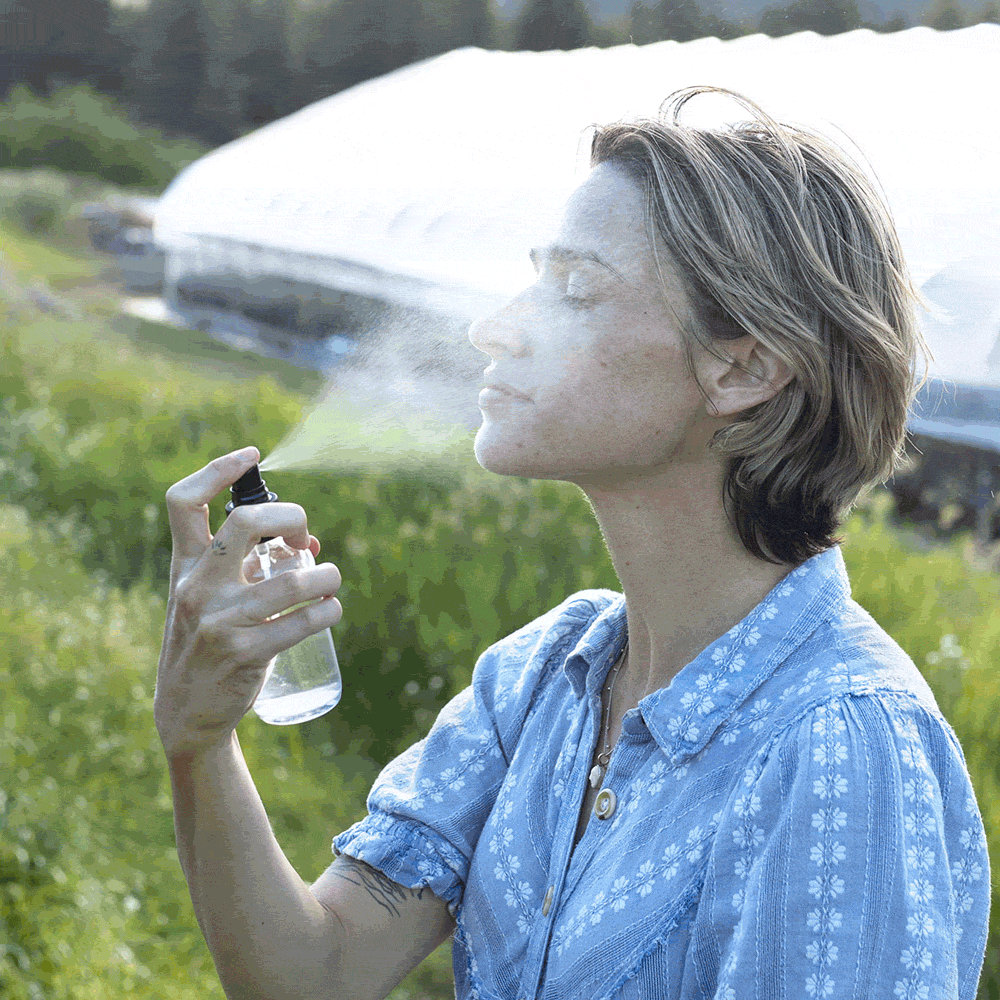 Woman sprays Rose Geranium HydroSoul on her face outdoors in a sunny garden