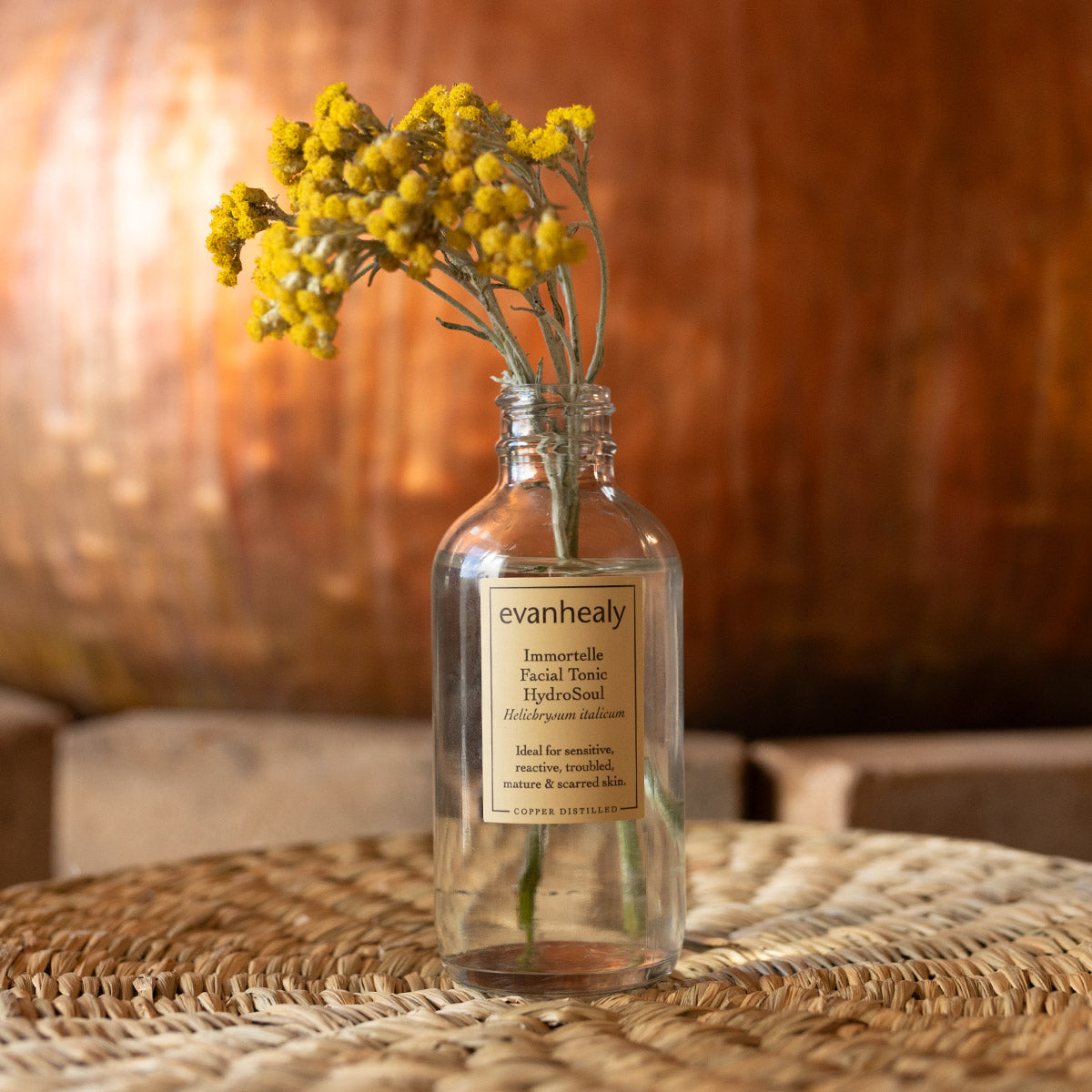 Front view of Immortelle Facial Tonic HydroSoul showcasing clear bottle and yellow flowers with warm wooden background