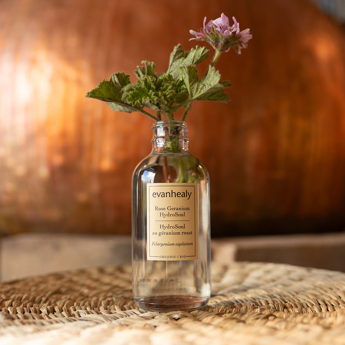 Front view of Rose Geranium HydroSoul bottle showcasing a geranium sprig on a woven surface against a warm wooden background