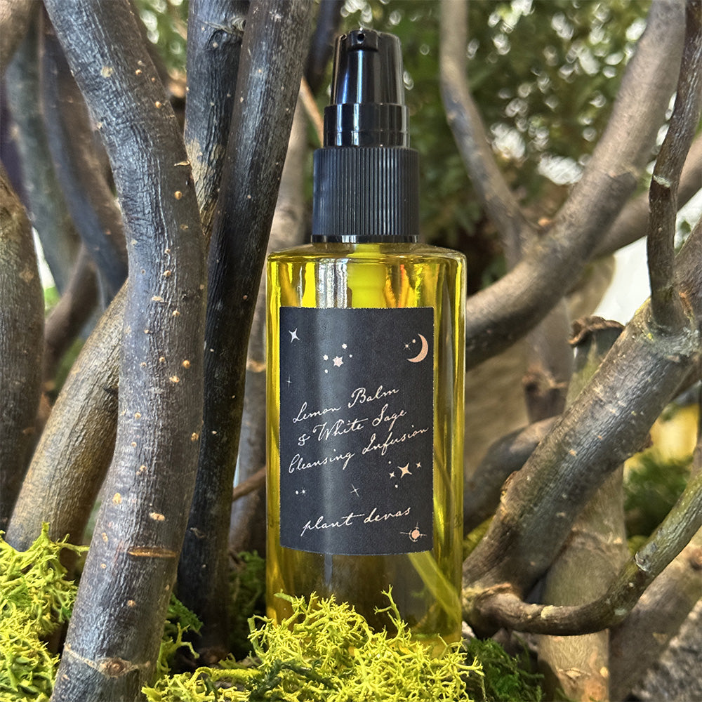 Front view of Lemon Balm & White Sage Cleansing Infusion pump bottle with golden oil and black label among branches and moss