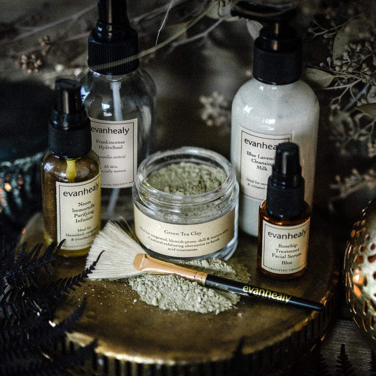 Assortment of evanhealy skincare products on a round wooden tray, an open jar of Green Tea Clay with a brush and dried flowers in moody lighting
