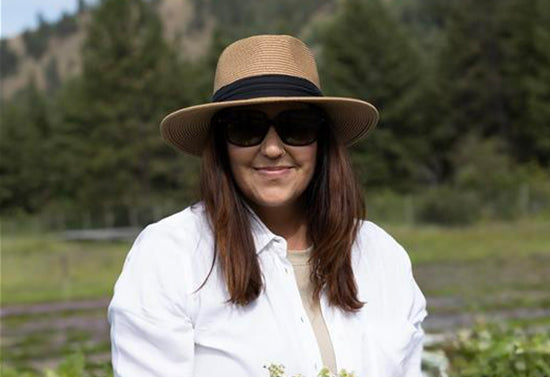Woman wearing a straw sun hat and sunglasses, smiling in a sunlit garden with trees in the background