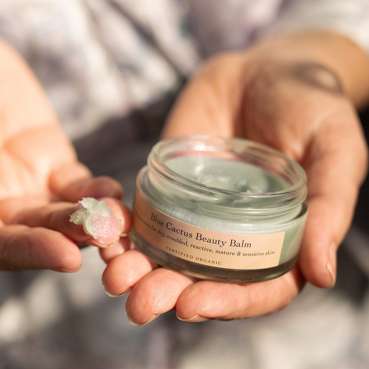 Close-up view of Blue Cactus Beauty Balm showcasing pale green balm on a fingertip and in an open jar with hands holding it
