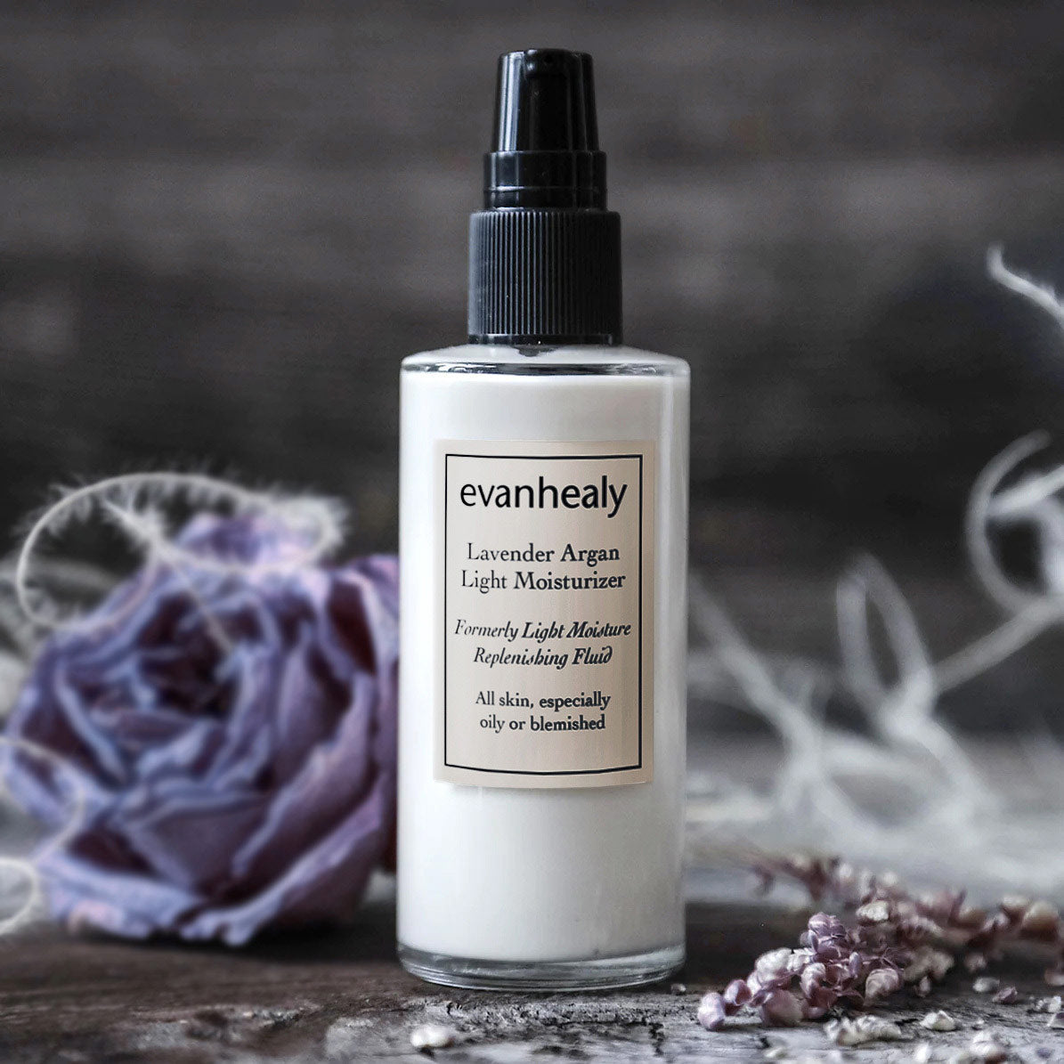 Front view of Lavender Argan Light Moisturizer pump bottle with lavender flowers and dried botanicals on a dark surface