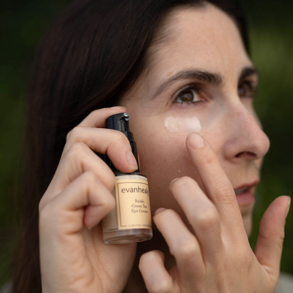 Woman applying Reishi Green Tea Eye Cream under her eye while holding the bottle