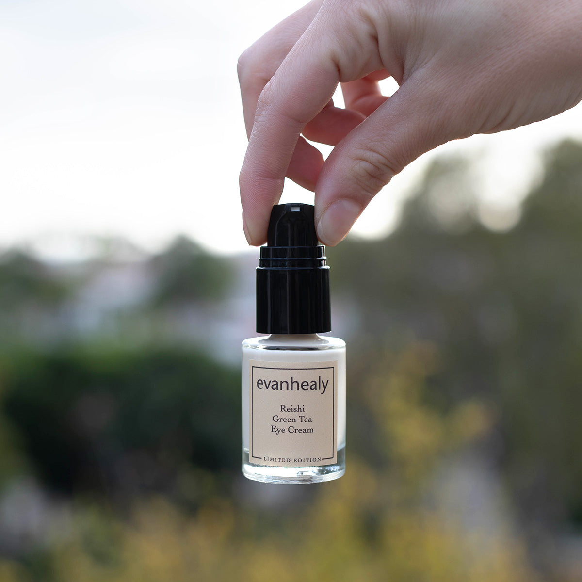 Close-up of Reishi Green Tea Eye Cream bottle with black pump held against a blurred outdoor background
