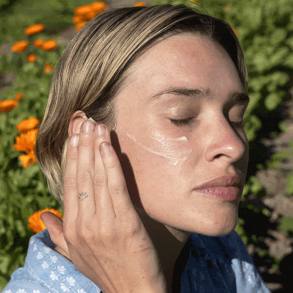 Close-up of a person gently applying whipped shea moisturizer to their cheek outdoors with orange flowers in the background