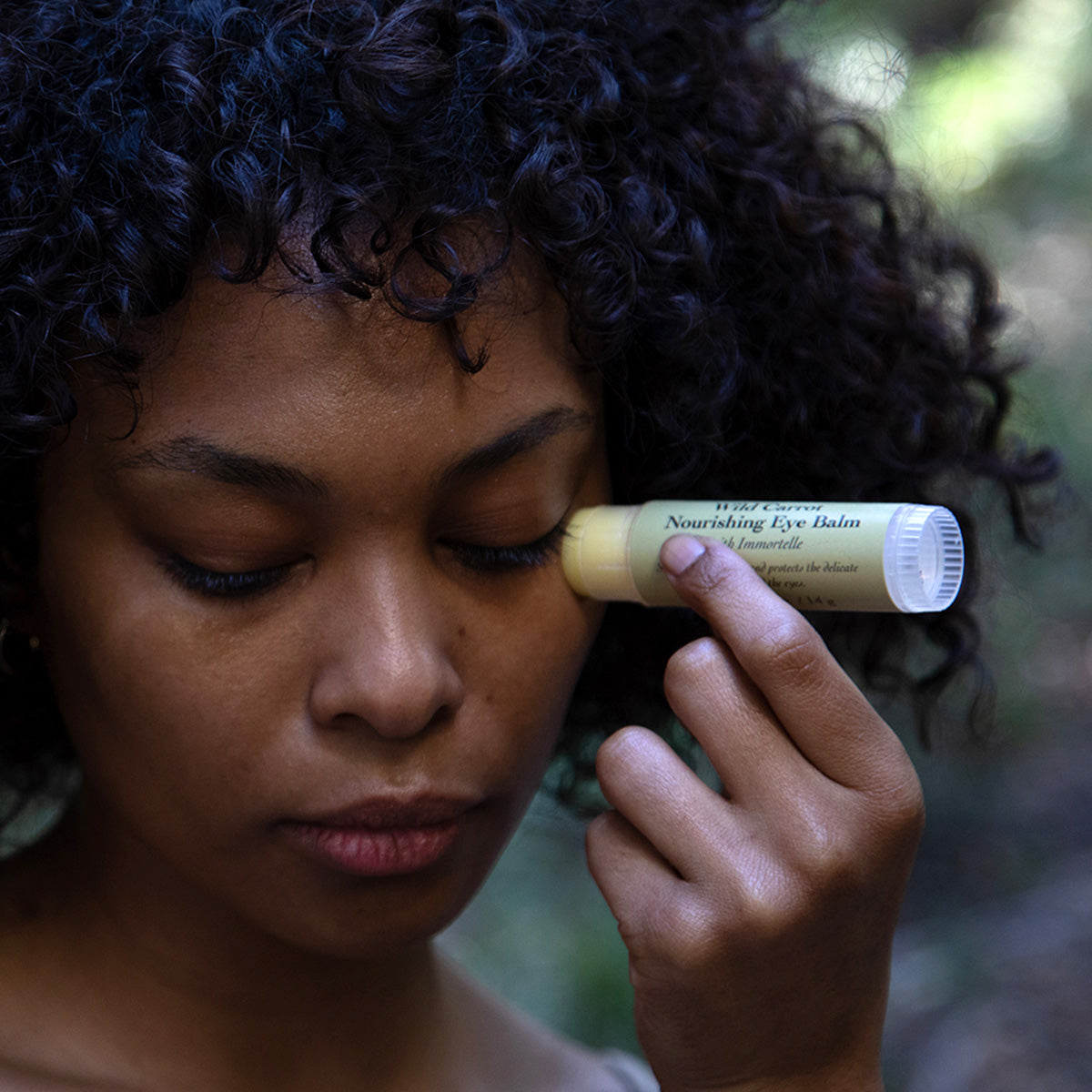 Person applying Wild Carrot Immortelle Eye Balm beneath the eye in a softly lit outdoor setting