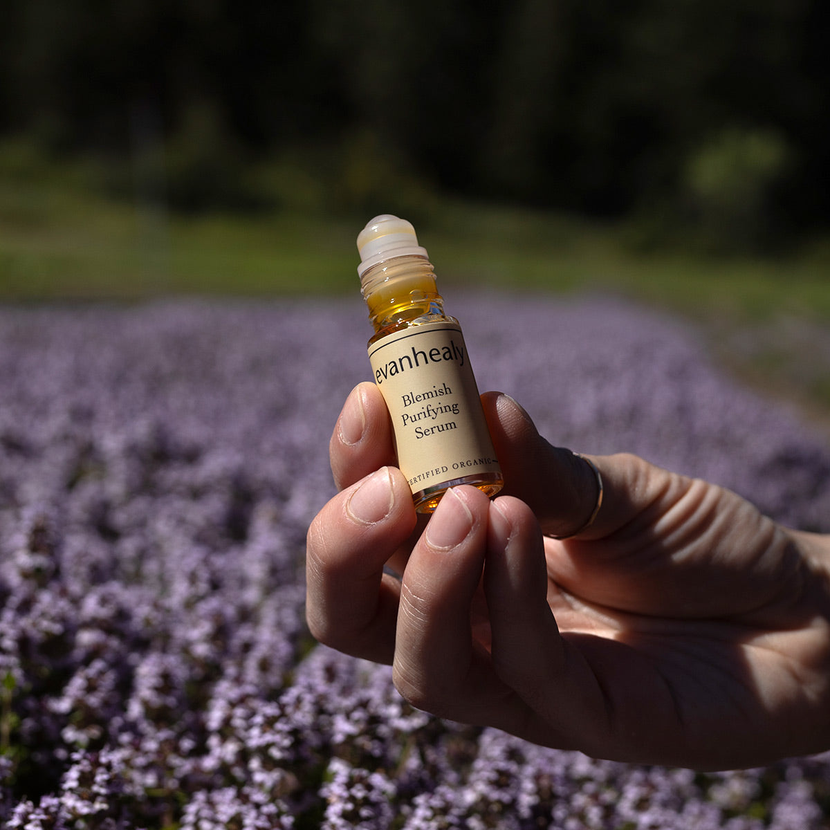 Close-up of evanhealy Blemish Purifying Serum showcasing its amber roll-on bottle with purple flowers in the background