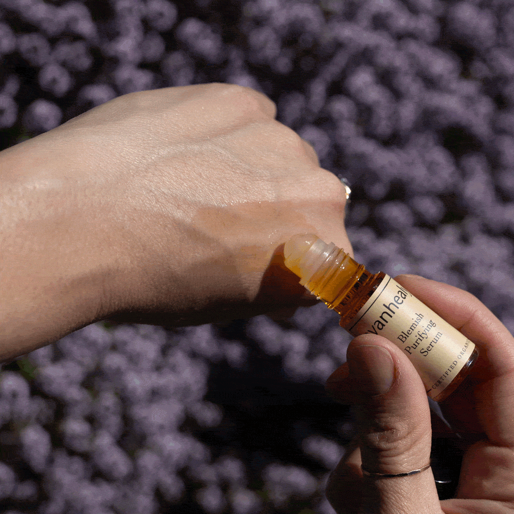 Close-up of evanhealy Blemish Purifying Serum roll-on bottle applying serum to a hand with purple flowers in background