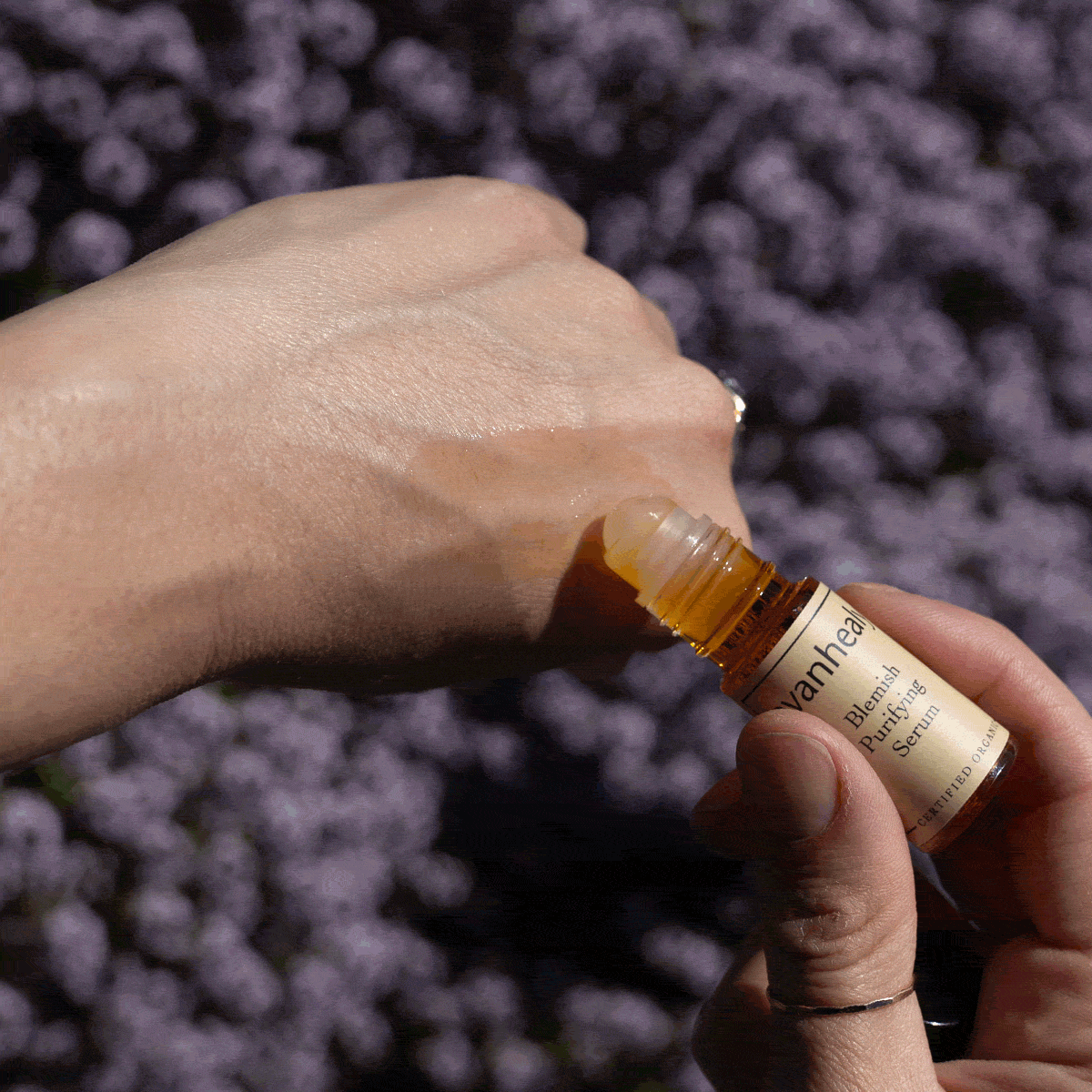 Close-up of evanhealy Blemish Purifying Serum roll-on bottle applying serum to a hand with purple flowers in background